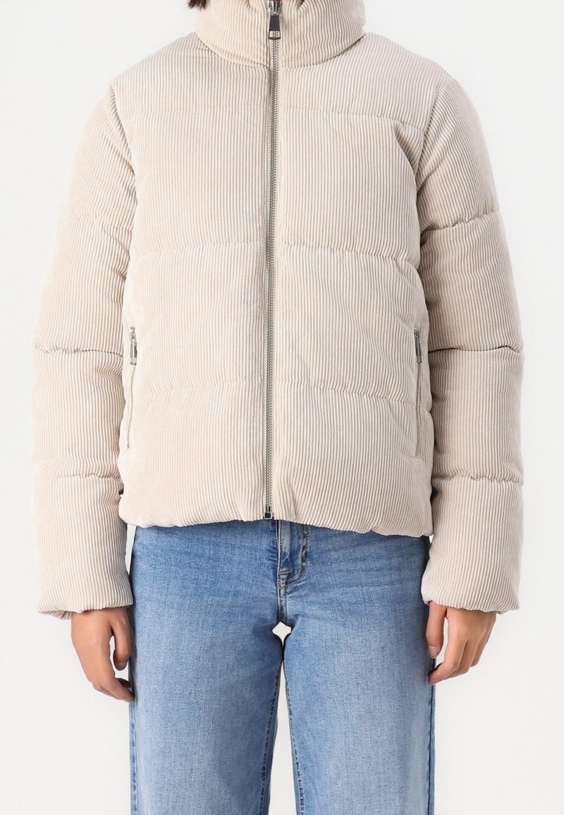 Personne portant une veste matelassée en velours côtelé beige avec fermeture éclair devant et un jean bleu clair, debout devant un fond uni.