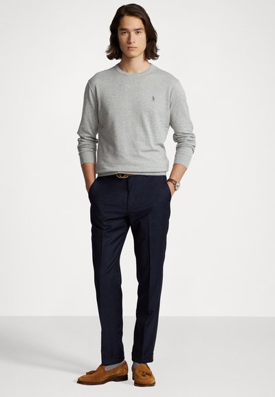 Šviesiai pilkas megztinis su apvalia iškirpte, tamsiai mėlyni kelnių, rudi zomšiniai loafers batai ir laikrodis. Modelis stovi su rankomis kišenėse.