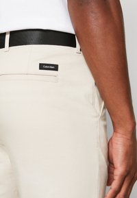 Beige Calvin Klein Hose mit schmaler Passform, mit schwarzem Bund und einem kleinen schwarzen Logoschild auf der Gesäßtasche.