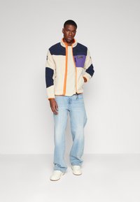 Fleece jacket in cream, navy, and purple with orange zipper. Features a zippered chest pocket and relaxed fit, paired with light blue jeans.