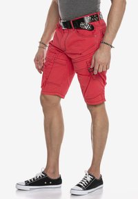 Rote Cargoshorts aus Baumwolle mit mehreren Seitentaschen, kontrastierenden Nähten und einem schwarz genietetem Gürtel. Getragen mit schwarzen Leinenschuhen.