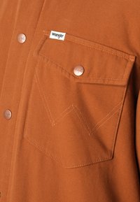 Bruine katoenen shirt met een knoopzak op de borst, voorzien van een Wrangler-logo. Koperen knopen en een kenmerkend gestikt patroon op de zak.
