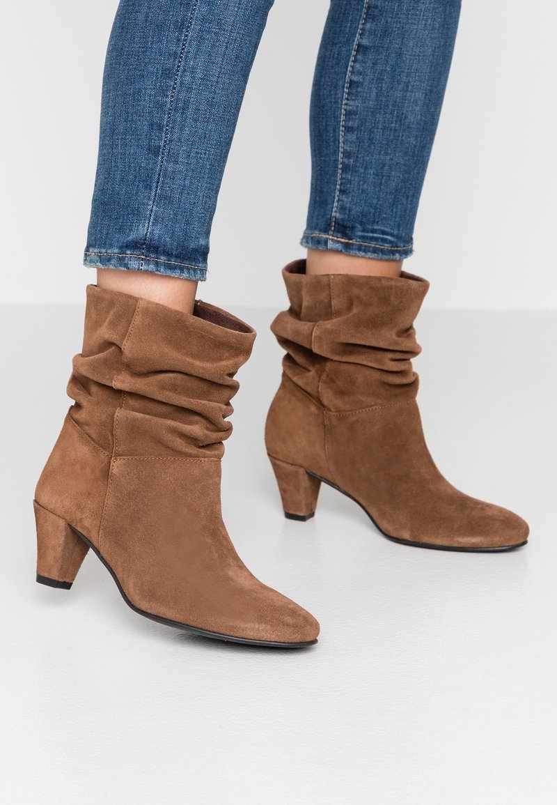 Bottines marron en daim ample avec talons blocs de hauteur moyenne, portées avec un jean skinny bleu sur un fond clair uni.