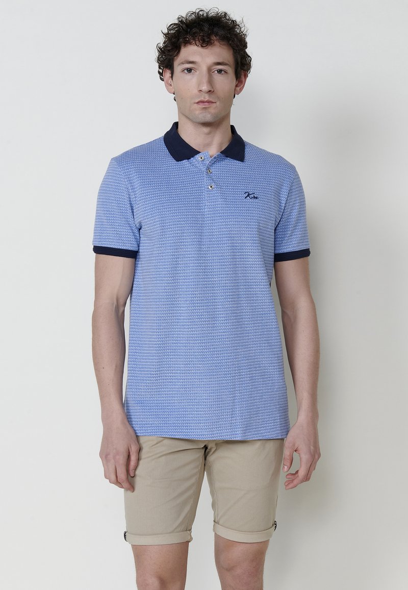 Gai zilas polo krekls ar teksturētu rakstu, tumši zila apkakle un piedurkņu aproces, saskaņots logo uz krūšu; kombinēts ar bēšiem salocītiem šortiem.