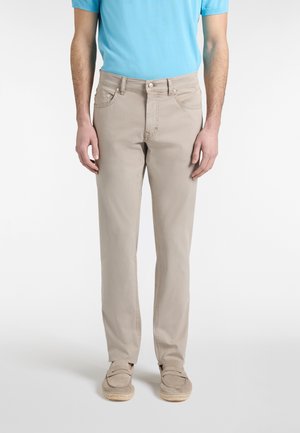 Uomo che indossa pantaloni beige a gamba dritta, camicia a maniche corte azzurro chiaro e scarpe slip-on beige, in piedi contro uno sfondo bianco.