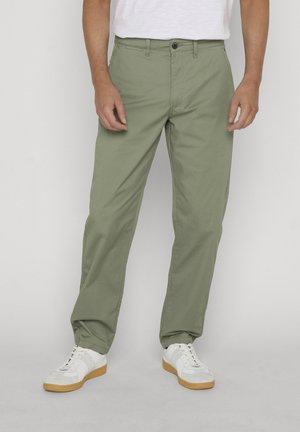 Uomo che indossa pantaloni dritti color verde oliva chiaro e sneakers bianche, in piedi davanti a uno sfondo semplice.