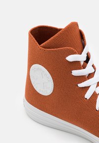 Sapatilha alta laranja com upper de tecido texturizado, sola de borracha branca, atacadores brancos e um patch de logotipo circular na lateral.