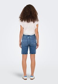 Vidēji zili džinsa šorti ar taisnu griezumu, saplēstu apakšmalu un klasiskām aizmugurējām kabatām, savienoti ar baltu ar volāniem rotātu blūzi.