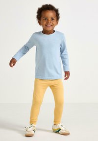 Smilende toddler med krøller iført en lyseblå langærmet skjorte, gule leggings og hvide sneakers med grønne og gule striber.