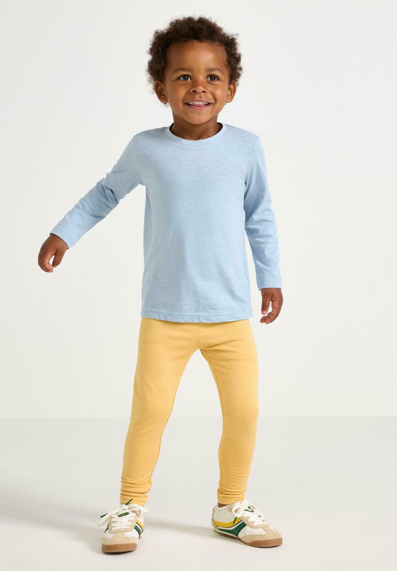 Smilende toddler med krøller iført en lyseblå langærmet skjorte, gule leggings og hvide sneakers med grønne og gule striber.