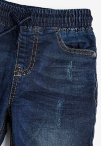 Gros plan d'un pantalon en denim bleu foncé avec taille élastique et cordon, avec poches avant et détails de coutures jaunes.