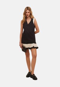 Top negru fără mâneci cu decolteu în V, asortat cu o fustă plisată crem și neagră și mocasini negri. Ținând o geantă de tip clutch neagră.