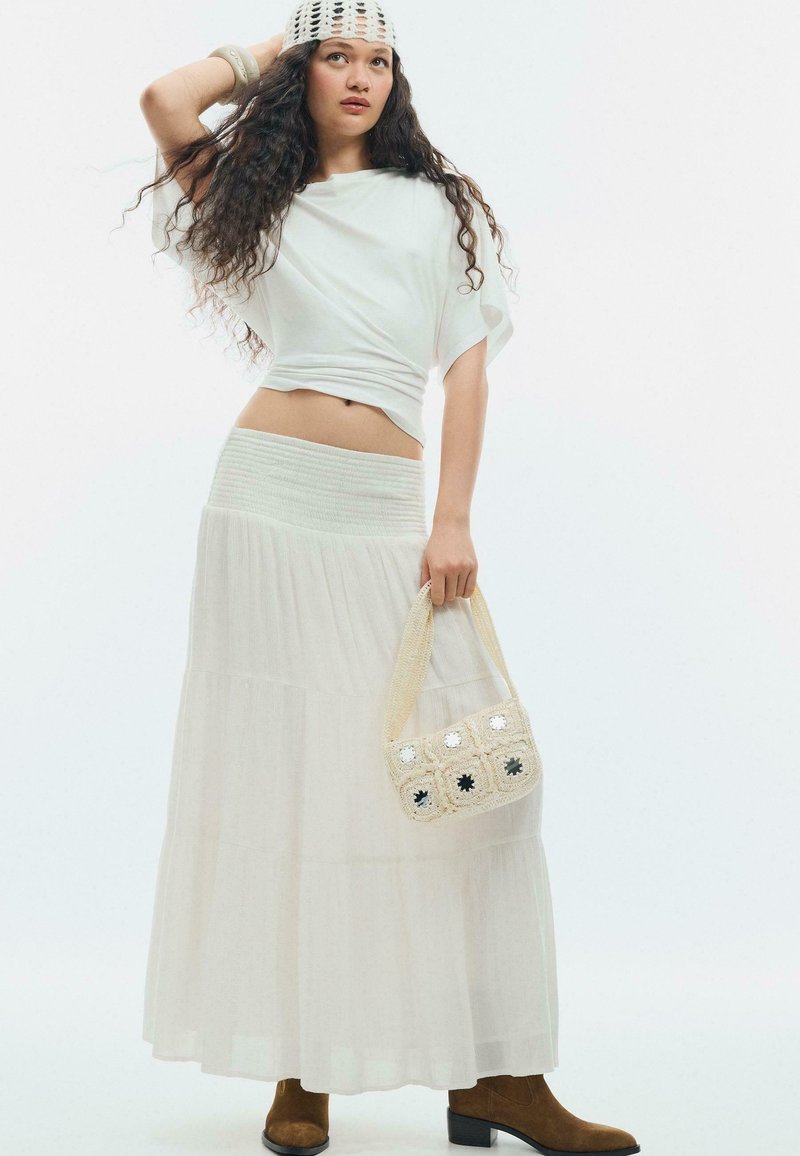 Femme aux longs cheveux bouclés portant un haut blanc, une longue jupe blanche, un chapeau au crochet, tenant un sac à main au crochet et des bottines marron.