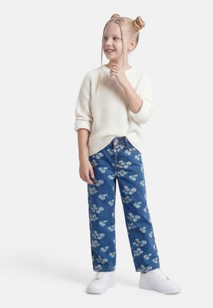 Niña joven con cabello rubio en trenzas y moños, vestida con un suéter blanco, vaqueros azules con flores y zapatillas blancas, de pie, sonriendo y mirando hacia la derecha.