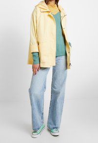 Gul vattentät jacka över en grön tröja och randig polotröja, parat med vida denimjeans och sneakers med gröna detaljer.