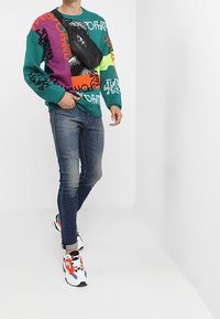 Grön och flerfärgad grafisk tröja med svart midjeväska, blå jeans och flerfärgade sneakers. Texturerat tyg och djärva mönster.