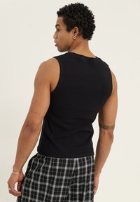 Svart ermeløs tanktopp med ribbestruktur, tettsittende design. Bæres med svart-og-hvit rutete shorts. Synlig tatovering på overarmen.