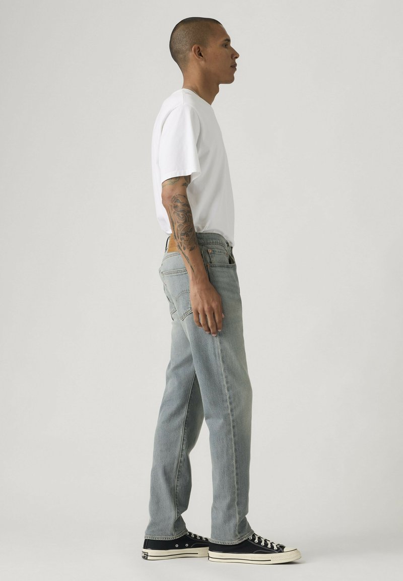 Homme à la tête rasée et tatouages sur les bras, portant un t-shirt blanc, un jean bleu clair et des baskets noires, debout de profil sur un fond uni.