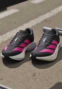 Ζευγάρι μαύρα και ροζ παπούτσια Adidas Adizero τοποθετημένα σε άσφαλτο κοντά σε λευκές γραμμές.
