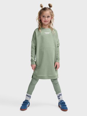 Junges Mädchen mit blondem Haar in zwei Dutts, trägt ein hellgrünes langes Sweatshirt, passende Leggings, weiße Socken und blaue Turnschuhe, steht breitbeinig.