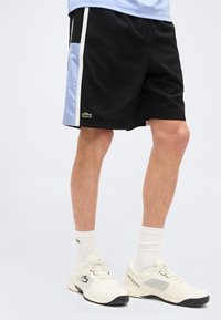 Πόδια με μαύρο σορτς με μπλε και λευκές ρίγες στο πλάι, λευκές κάλτσες μέσης και λευκά παπούτσια Lacoste με μαύρες σόλες.