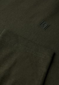 Camiseta de color verde oliva con una textura suave, que presenta un bolsillo en el pecho y un pequeño logo negro en la esquina superior derecha.