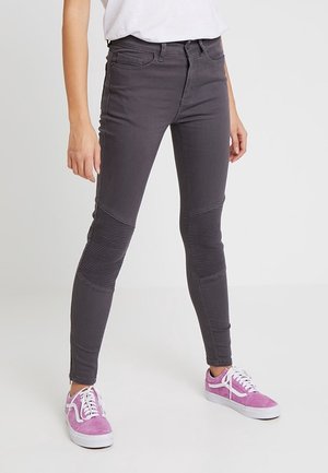 Personne portant un pantalon ajusté gris foncé avec des panneaux côtelés aux genoux et des baskets roses avec des lacets et semelles blanches, debout sur un sol clair.
