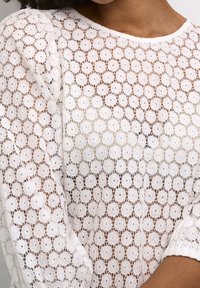 Haut en dentelle blanche avec un motif floral à finition texturée. Le design comprend des manches longues et un col rond.