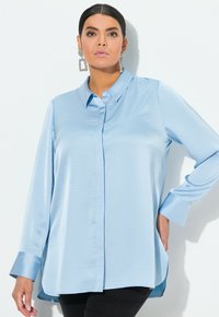 Blusa di raso blu chiaro con chiusura a bottoni, maniche lunghe e orlo leggermente curvo. Finitura testurizzata e polsini a contrasto.