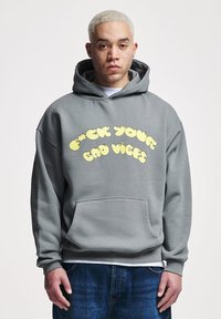 Felpa grigia in cotone, con tasca frontale e grande scritta grafica gialla che dice "F*CK YOUR BAD VIBES." Vestibilità standard con polsini a coste.