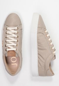 Baskets basses beige avec semelles blanches, lacets crème passant par des œillets dorés, tissu à motif subtil, et détail de semelle intérieure de marque.