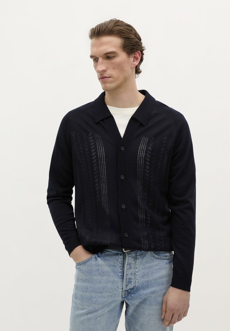 Cardigan tricotat bleumarin cu un model vertical texturat, cinci butoane și un guler plat. Purtați peste o cămașă albă și asortați-l cu blugi deschiși.