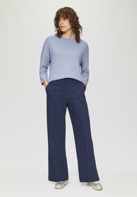 Vrouw met krullend haar, gekleed in een lichtblauwe trui, marineblauwe brede broek en zilveren sneakers, staand met haar handen in de zakken.