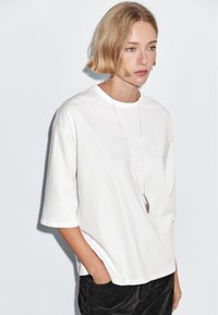 Woman with short blonde hair wears oversized white t-shirt, long silver necklace, and black pants, standing against plain light background.