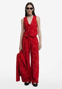Rood, gebloemd vest en wijde broek met een bijpassende jas. Model draagt zwarte zonnebril en een choker ketting.