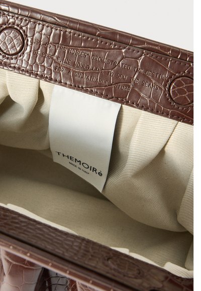 Dentro de uma bolsa de couro com textura de crocodilo castanho, forrada com tecido bege e com um rótulo branco com a inscrição "THEMOIRé MADE IN ITALY."