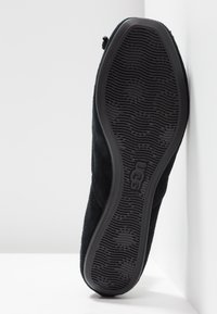 Černá nubuková nazouvací bota s texturovanou černou gumovou podrážkou, která má vlnkový vzor a malé vyražené logo UGG na podrážce.