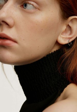 Prenda negra de cuello alto con textura acanalada, diseño ajustado. Aro metálico en la oreja. Piel con pecas y cabello rojizo visibles.