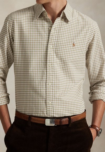 Bărbat purtând o cămașă bej și albă în carouri cu nasturi, mâneci suflate, curea maro, pantaloni închiși la culoare și ceas la încheietura stângă.