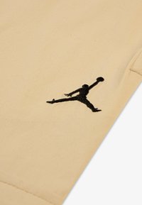 Beige stoff med en svart brodert Air Jordan-logo. Materialet ser mykt ut med en jevn tekstur.