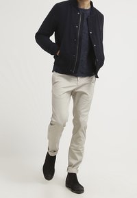 Uomo che cammina indossando una giacca bomber blu navy, un maglione blu scuro, pantaloni beige chiaro e stivali neri alla caviglia su uno sfondo bianco.