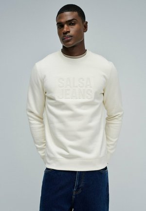 Felpa color crema con colletto e polsini a costine, con testo in rilievo "SALSA JEANS" sul davanti. Indossata con jeans blu scuro.