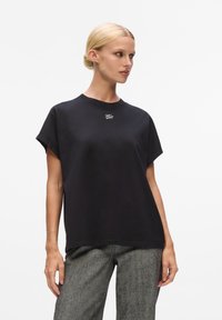 Schwarzes Baumwoll-oversized T-Shirt mit kurzen Ärmeln, versehen mit einem dezenten weißen Logo auf der Brust, kombiniert mit grauen, strukturierten weit geschnittenen Hosen.