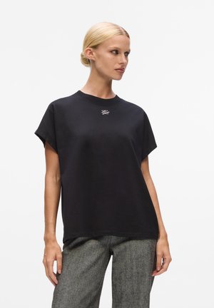 Black cotton oversized t-shirt with short sleeves, features a subtle white logo on the chest, paired with gray textured wide-leg trousers.