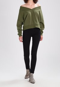 Olivgrön off-shoulder tröja med V-ringning, sänkt axel och ribbad fåll, kombinerad med svarta tajta byxor och ankelboots i leopardmönster.