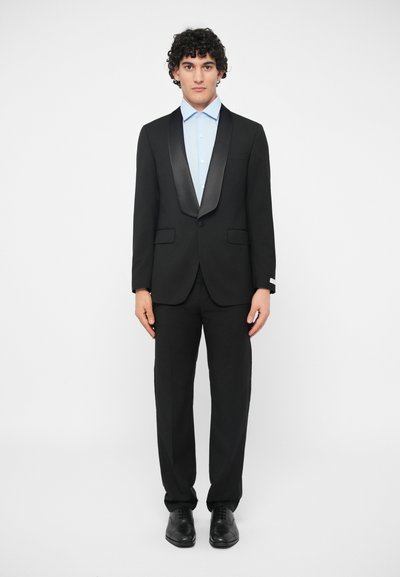 Black suit with satin lapels, paired with a light blue shirt. Structured fit, featuring two pockets on the jacket and black dress shoes.