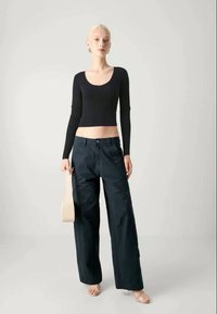 Top cropped preto de mangas longas com um decote redondo, combinado com calças largas azul-escuras; modelo segura uma mala bege. Fundo neutro.