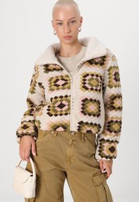 Kort jacka i krämfärgad fleece med geometriska mönster i brunt, grönt och rosa. Dragkedjestängning och en mjuk krage. Matchas med khakifärgade byxor.
