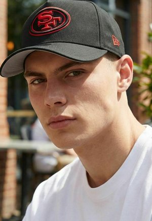 Junger Mann mit schwarzer San Francisco 49ers Mütze und weißem Hemd, der in einer Outdoor-Umgebung mit verschwommenem Hintergrund geradeaus schaut.