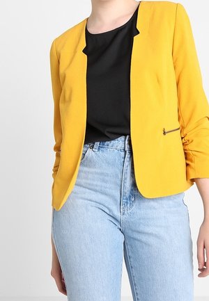 Person wearing a bright yellow blazer, black top, and high-waisted light blue jeans against a plain white background.